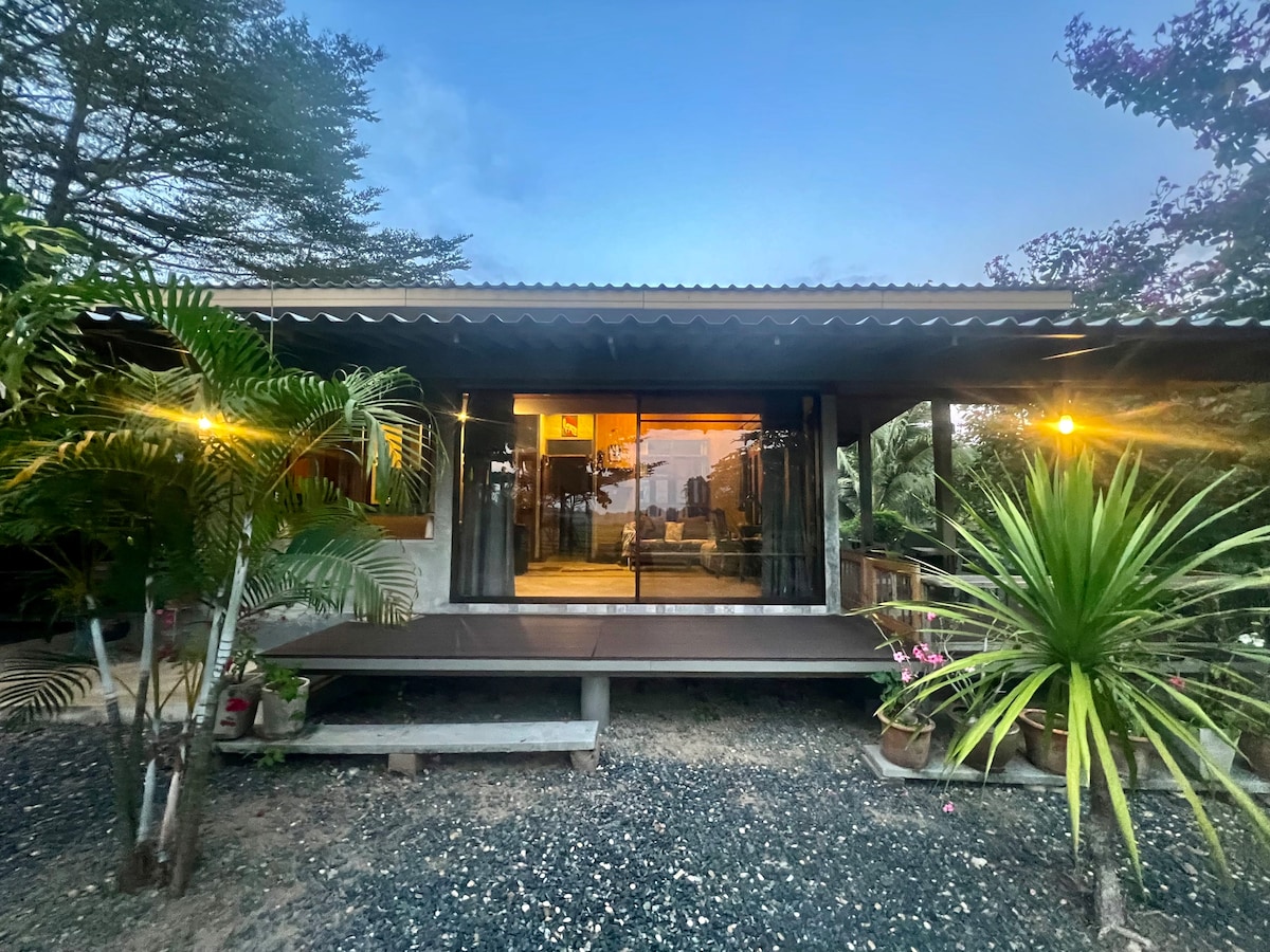 The bungalow is framed by greenery and positioned under a gentle evening sky. Its front is glass-paneled, allowing natural light to fill the interior. Potted plants are placed along the gravel pathway, enhancing the welcoming entrance.