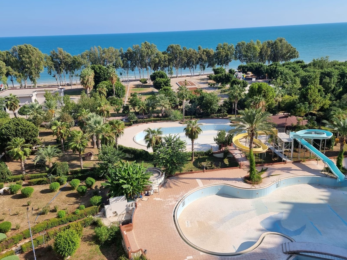 A scenic view captures a landscaped area with well-maintained gardens and palm trees overlooking a beach. A large swimming pool is featured, alongside water slides, surrounded by walking paths and green spaces, emphasizing a tranquil outdoor environment.