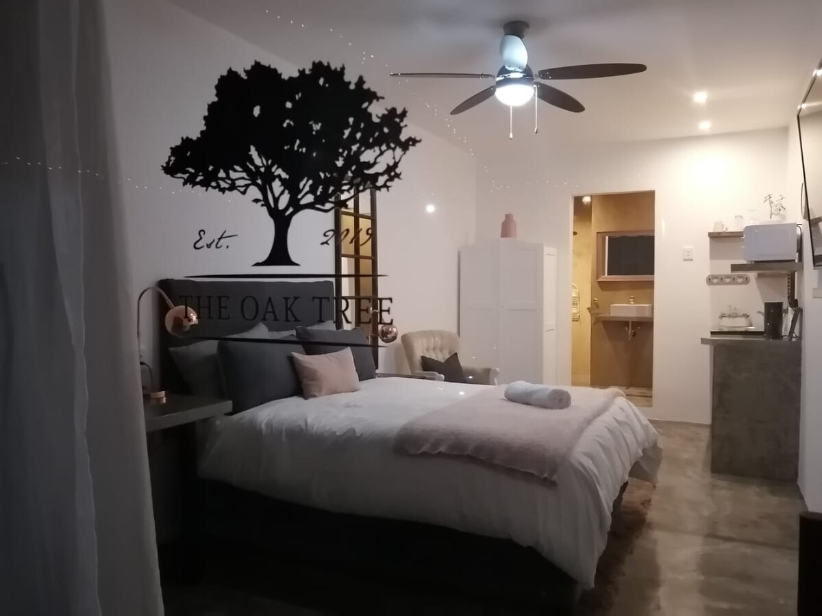 A well-lit, modern room features a spacious bed with neutral bedding. A logo of an oak tree is displayed on the wall. A comfortable chair and small table are positioned nearby. Soft lighting enhances the inviting atmosphere, while a ceiling fan provides additional comfort.