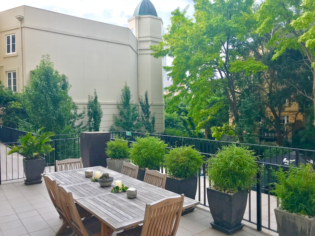 An expansive outdoor terrace features a large wooden dining table surrounded by comfortable seating. Lush greenery from potted plants adds a natural touch, while trees in the background create a serene setting. A crisp, open space invites moments of relaxation and dining al fresco.