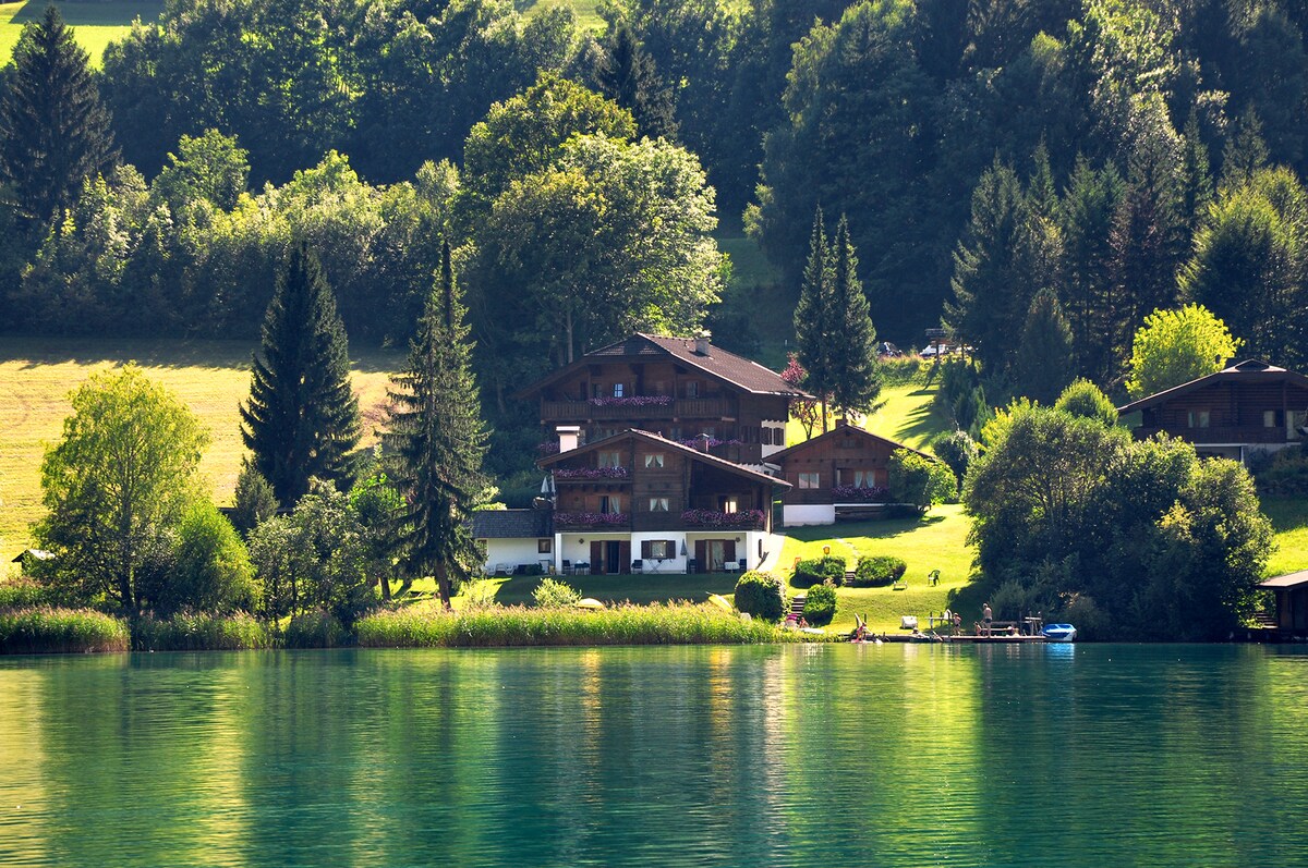 A scenic view features chalet-style buildings surrounded by lush greenery. The structures are set against a backdrop of trees and meadows, with their reflections visible on the calm lake water. The tranquil atmosphere emphasizes the natural beauty of the location.
