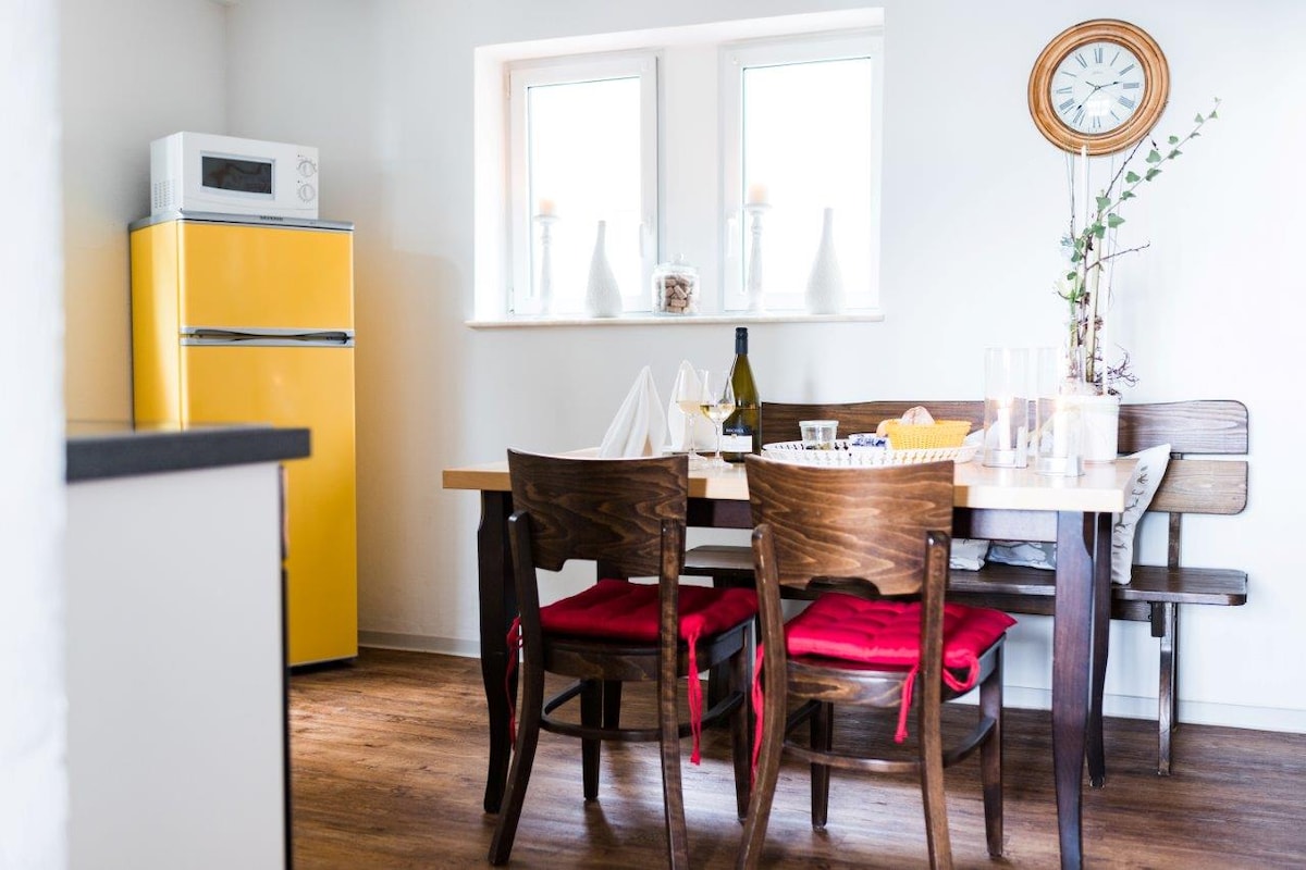 A bright kitchen area is showcased, featuring a yellow refrigerator and a wooden dining table surrounded by chairs with red cushions. Two windows allow natural light to fill the space, while decorative items are placed on the table, enhancing the inviting atmosphere.