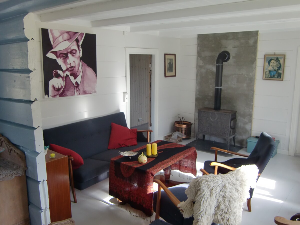 A comfortable seating area is presented with a dark sofa and armchairs, complemented by a wooden coffee table. Decorative items, including candles and a cozy throw, add character. A wood-burning stove is situated against a gray stone wall, enhancing the rustic feel of the space.