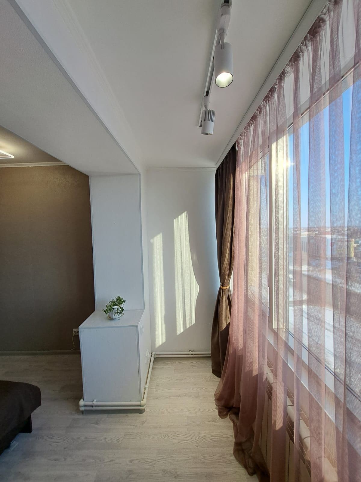 A well-lit corner of a room features tall windows draped with sheer and dark curtains. Natural light casts gentle shadows on the light-colored walls. A small decorative plant is positioned on a side table adjacent to a spacious bed with a simple design.