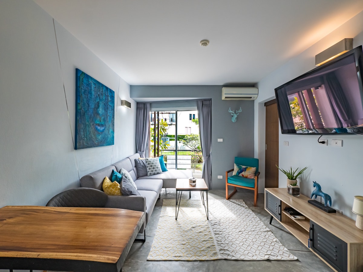 A modern living area features a light gray sofa with colorful accent pillows and a wooden dining table. A large flat-screen TV is mounted on the wall. Natural light enters through a sliding glass door, with greenery visible outside.
