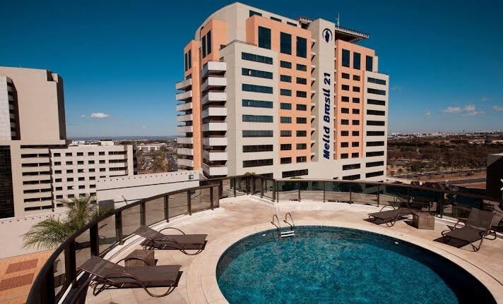 A rooftop pool area is depicted, featuring a circular swimming pool surrounded by poolside loungers. The building structure offers a modern design, with multiple floors visible in the background against a clear blue sky.