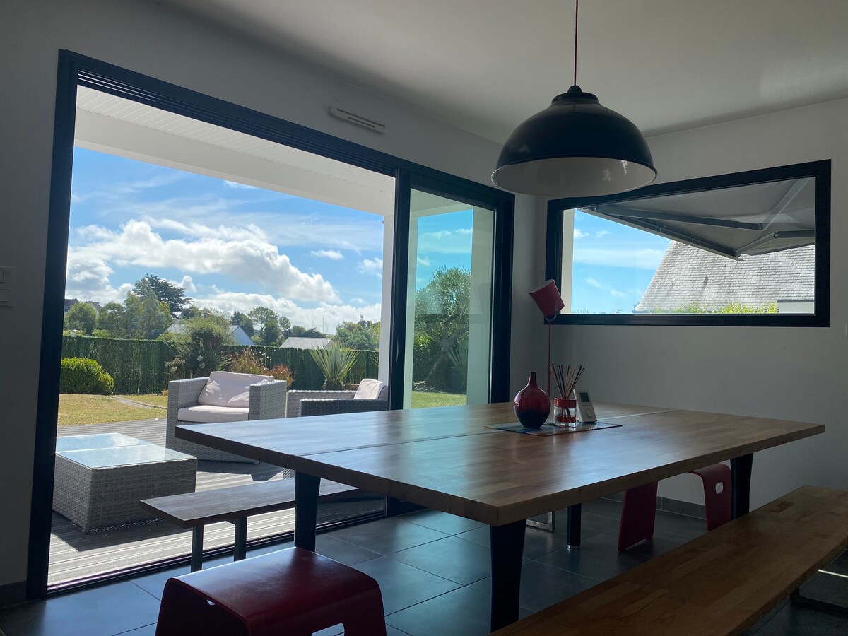 A spacious dining area features a large wooden table surrounded by red chairs. Large glass doors open to reveal a view of the garden and the outdoor seating area, allowing natural light to fill the space. A modern pendant light hangs above the table.