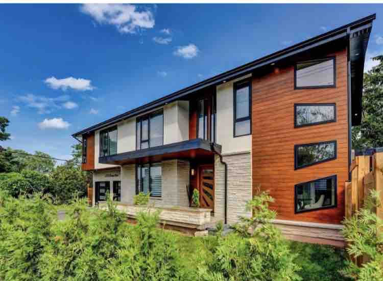A modern detached house features a blend of wood and stone exteriors, with large glass windows reflecting sunlight. The well-kept landscaping includes mature greenery, surrounding a spacious entrance and inviting front porch.