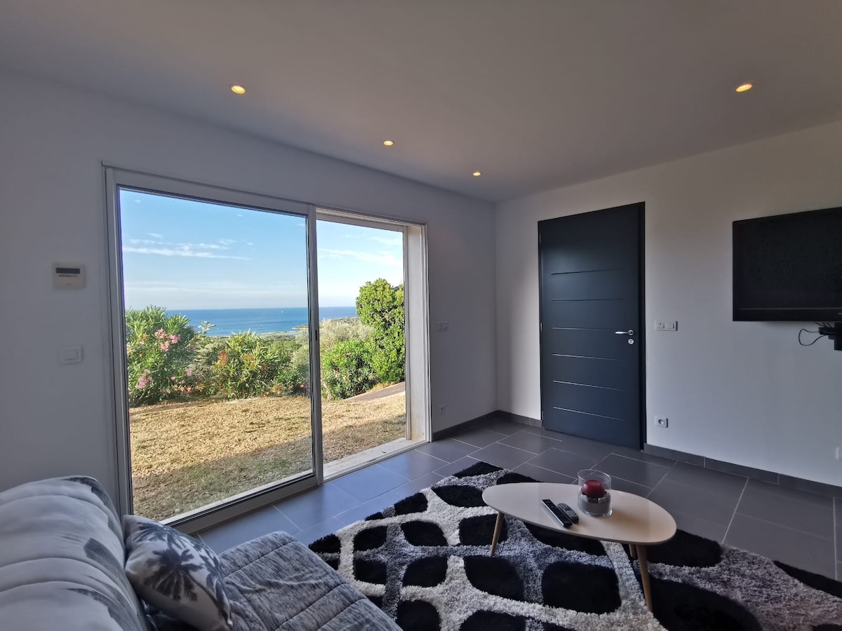 A modern living area is presented with a comfortable sofa and a low coffee table. A large sliding glass door opens to a view of greenery and the sea, allowing natural light to fill the space. A wall-mounted television is also visible.