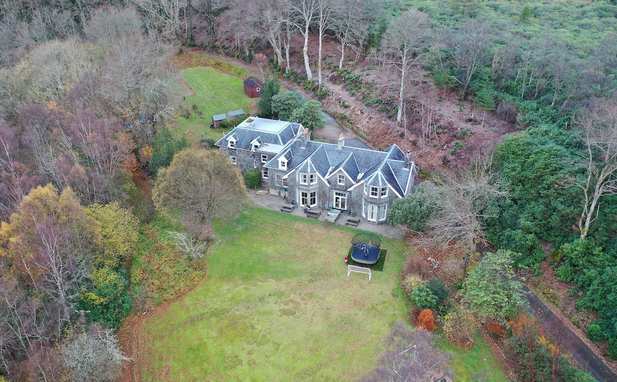 The aerial view showcases Ardvullin's spacious grounds, surrounded by diverse woodland. The restored cottage features a combination of traditional and modern architecture, with large windows reflecting the natural light. The expansive lawn is complemented by colorful vegetation, creating a serene outdoor setting.