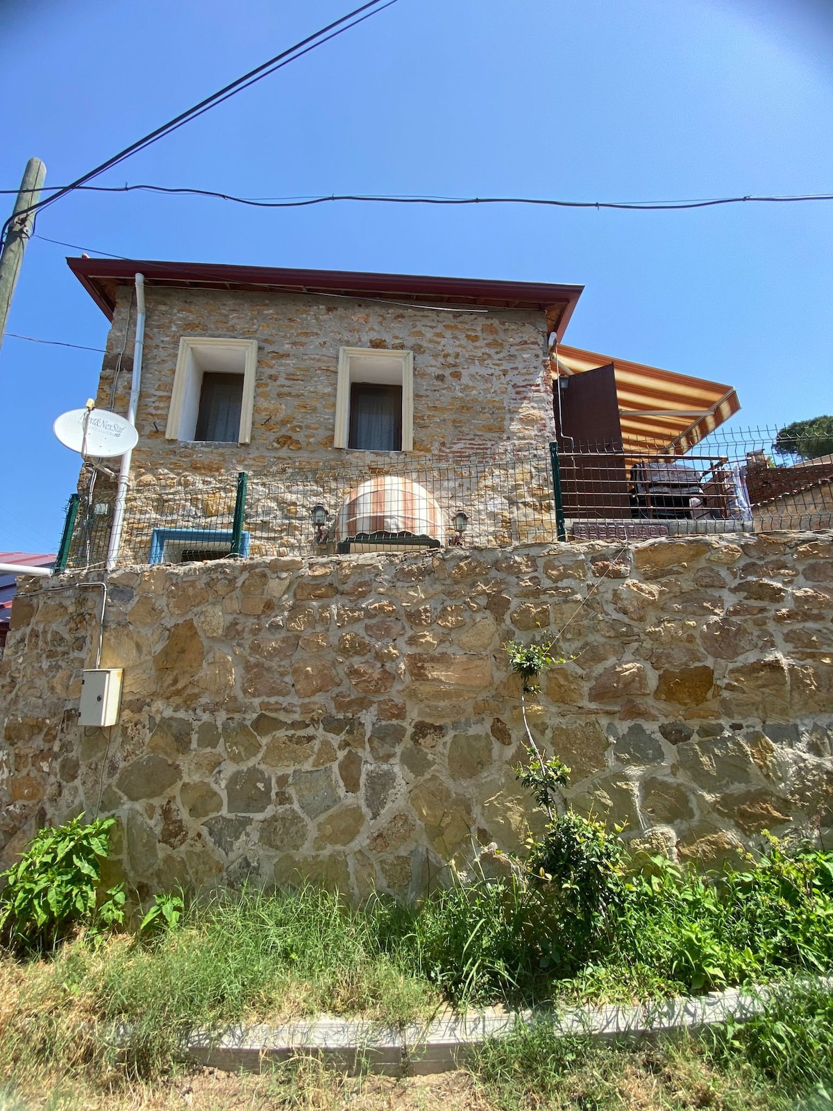 A rustic stone house with two levels is set against a clear blue sky. The structure features large windows and a striped awning, adding to its charm. A fence surrounds the property, with greenery visible in the foreground.