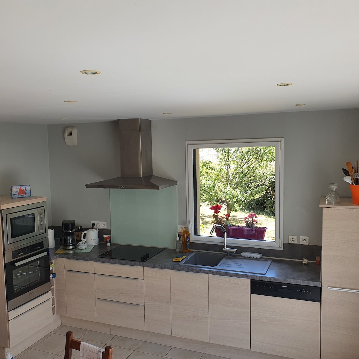A modern kitchen is presented with light wood cabinetry and a grey countertop. Stainless steel appliances, including an oven and coffee maker, are positioned neatly. A large window offers a view of greenery outside, allowing natural light to brighten the space.