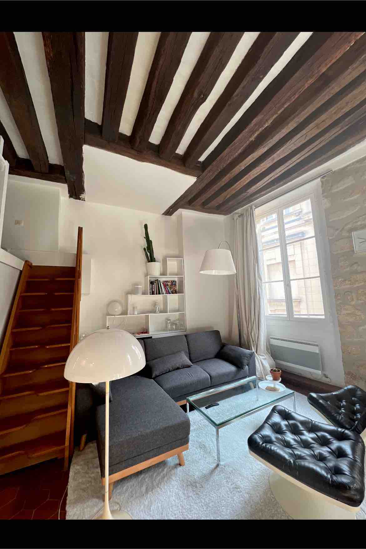 A living area features a dark sofa accompanied by a sleek glass coffee table, with natural light streaming through large windows. Exposed wooden beams add character to the ceiling, while a staircase leads to the mezzanine. A floor lamp and decorative items complete the cozy setting.