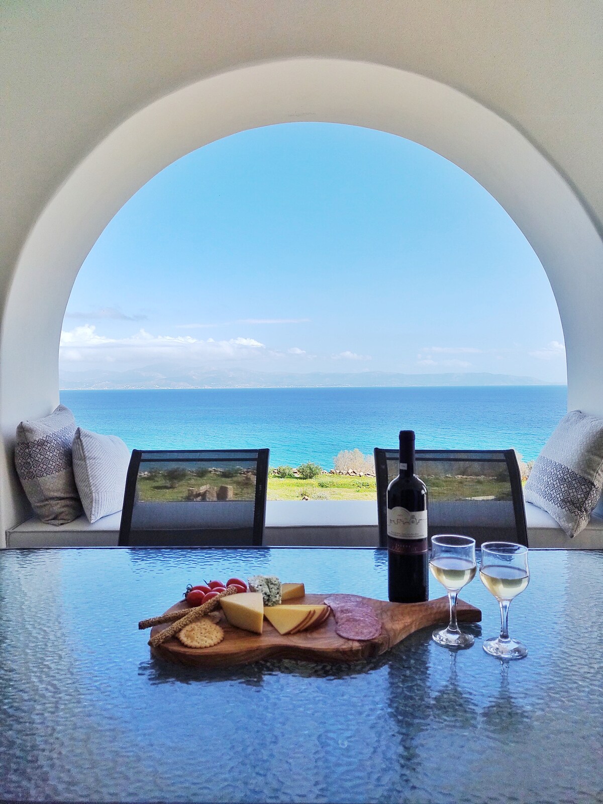 A scenic view is framed by a large arched window, showcasing an expansive sea and sky. A dining table is set with a wooden platter featuring an assortment of cheeses, cured meats, and colorful tomatoes. Wine is placed prominently behind the platter, inviting relaxation.
