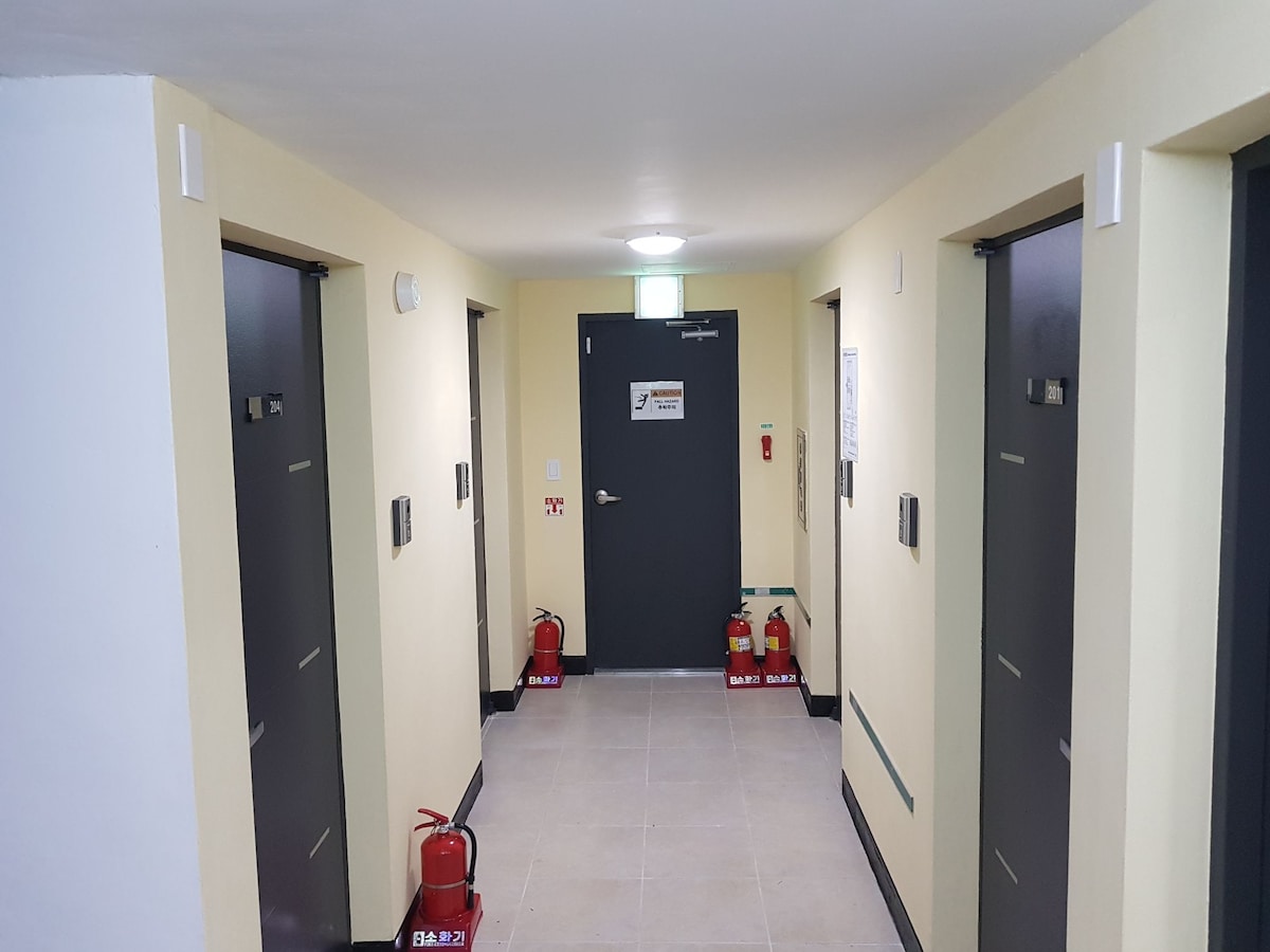 A well-lit hallway features multiple doors leading to guest accommodations, with fire extinguishers positioned along the walls for safety. Neutral-colored walls emphasize the space's clean design, and signs are displayed near the entrance to enhance navigational clarity.