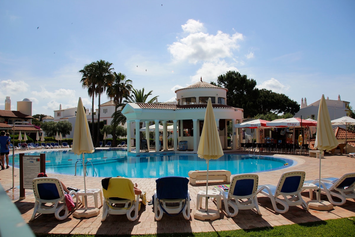A large outdoor pool is surrounded by sun loungers, some draped with colorful towels. Shade is provided by umbrellas, while nearby seating areas with tables are visible. The scene is complemented by palm trees and a building with a domed roof in the background.