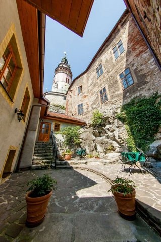 An inviting courtyard is framed by stone buildings, with a partially visible tower in the background. Green plants are placed in terracotta pots along the stone pathway, while a small table and chairs are situated under a clear blue sky.