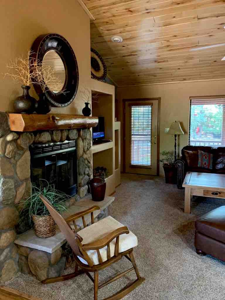 A cozy living area features a stone fireplace, contributing to a warm ambiance. A rocking chair is positioned nearby, and natural light filters through the window adorned with blinds. Decorative items, including plants and an accent mirror, enhance the inviting space.
