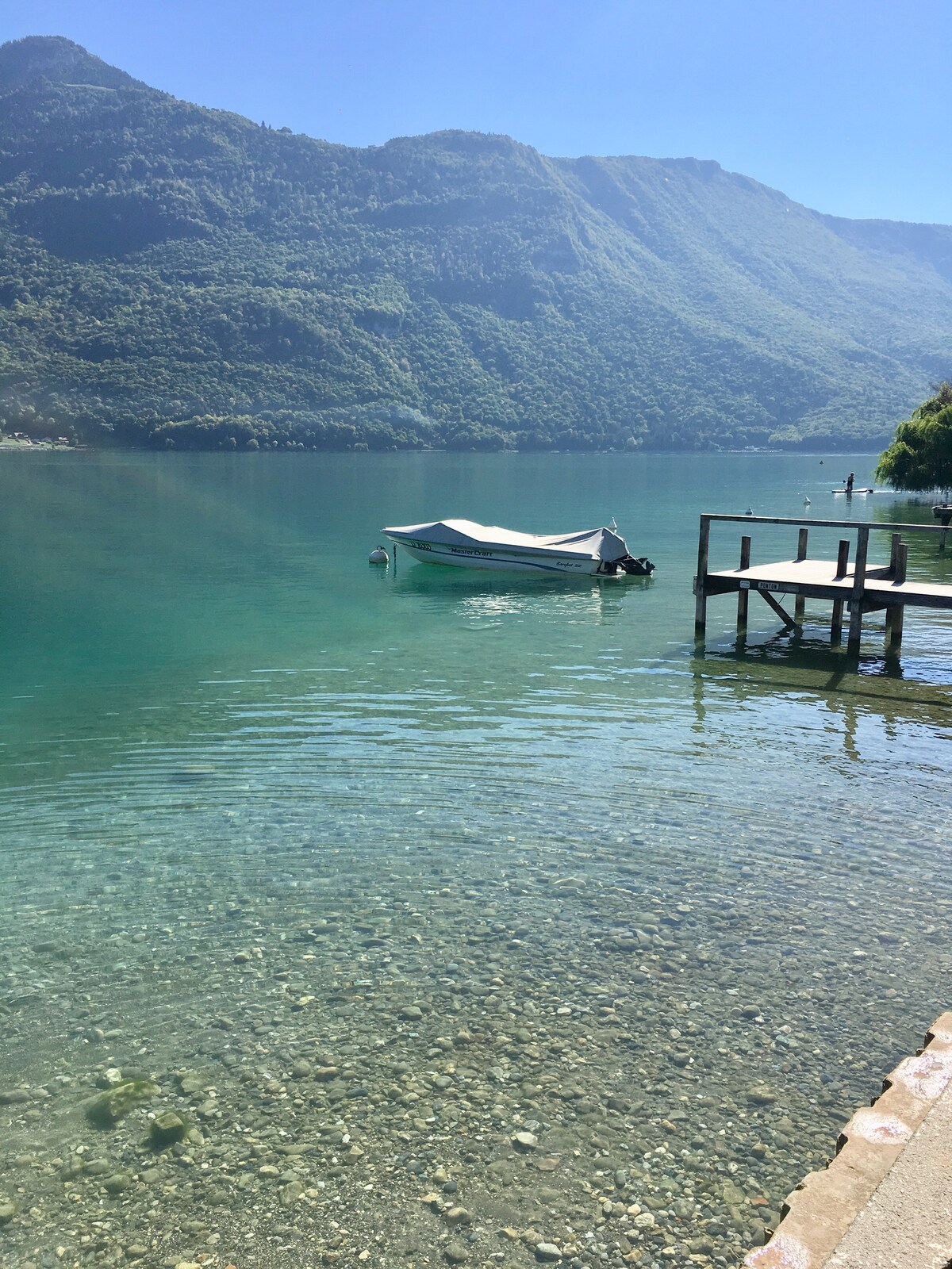 A clear lake is bordered by vibrant green mountains, reflecting the sunlight. A small boat is anchored nearby, with a wooden dock visible at the edge of the water. The lake's pristine surface reveals smooth stones beneath, creating a serene natural setting.