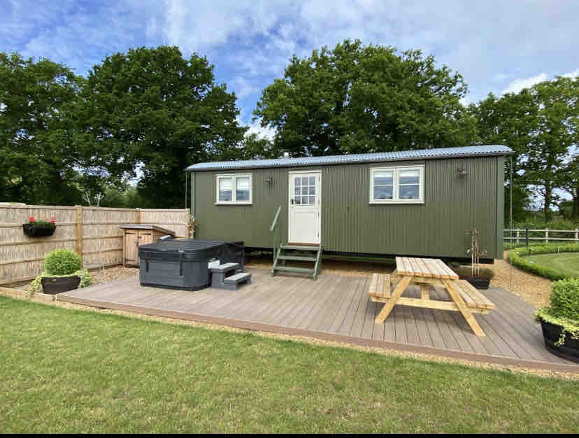 A shepherd's hut is set on a spacious deck with a hot tub and picnic table. Lush green grass surrounds the area, with wooden fencing and potted plants adding a touch of decoration. A wooden staircase leads to the entrance, framed by trees in the background.
