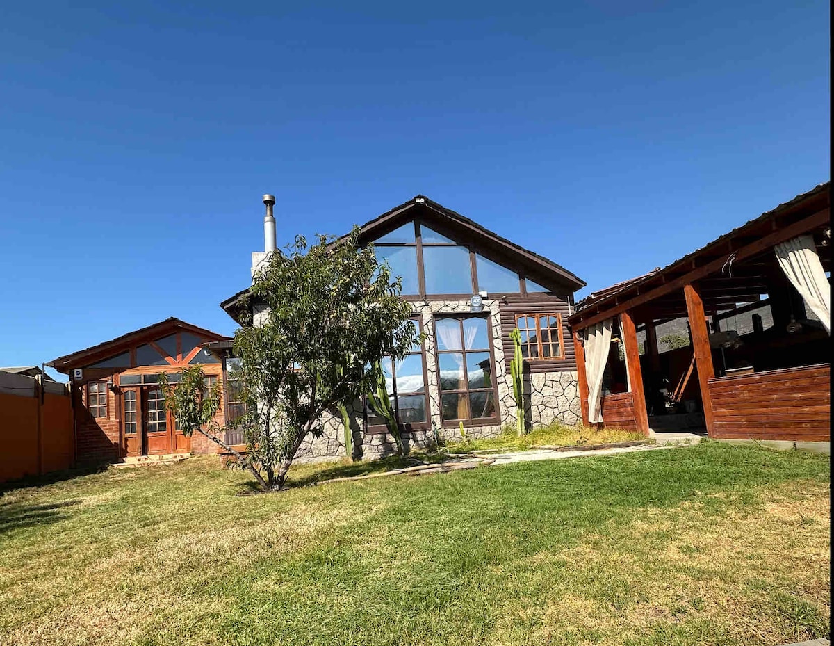 A rustic house features a stone and wooden façade under a clear blue sky. Large windows allow natural light to fill the interior. A grassy area surrounds the house, complemented by a separate outdoor structure with seating and draped fabric.