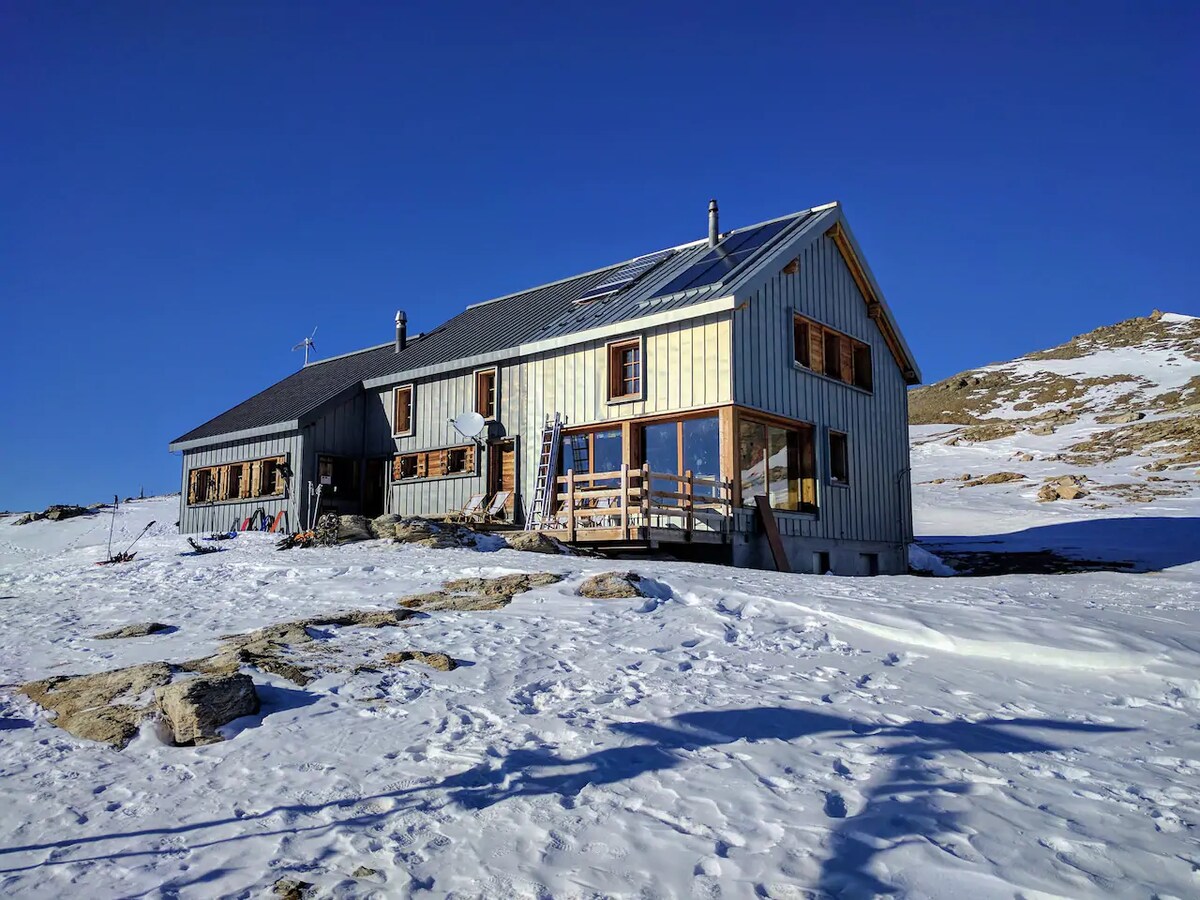 A mountain lodge is situated in a snowy landscape, featuring a combination of wood and metal siding. Solar panels are visible on the roof. Large windows allow for ample natural light, and a wooden deck extends from the building, overlooking the surrounding terrain.