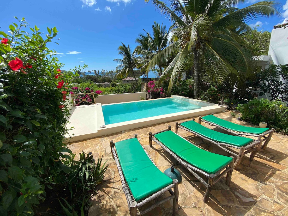 An outdoor area is depicted featuring a clear swimming pool surrounded by lush greenery and colorful flowers. Four green loungers are positioned beside the pool, allowing for relaxation in a tropical setting under the blue sky and palm trees.