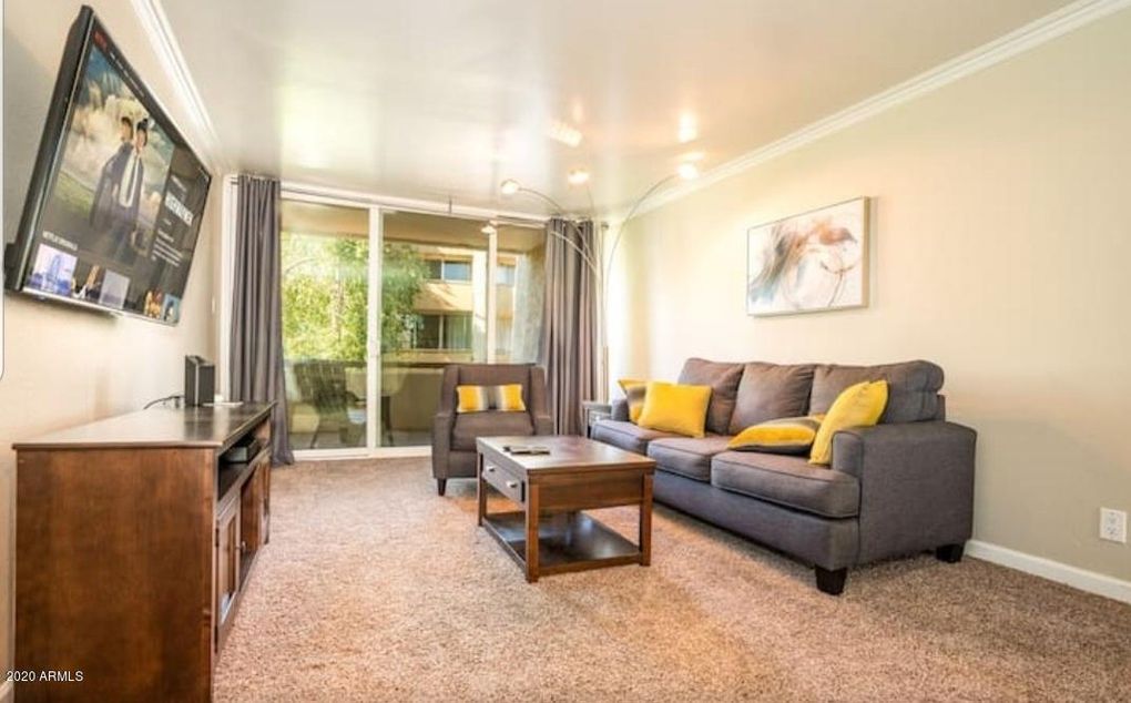 A cozy living area features a gray sofa with yellow accent pillows, complemented by a matching chair. A wooden coffee table sits in front, and a media unit is positioned against the wall. Large sliding glass doors allow natural light and provide access to the private balcony.
