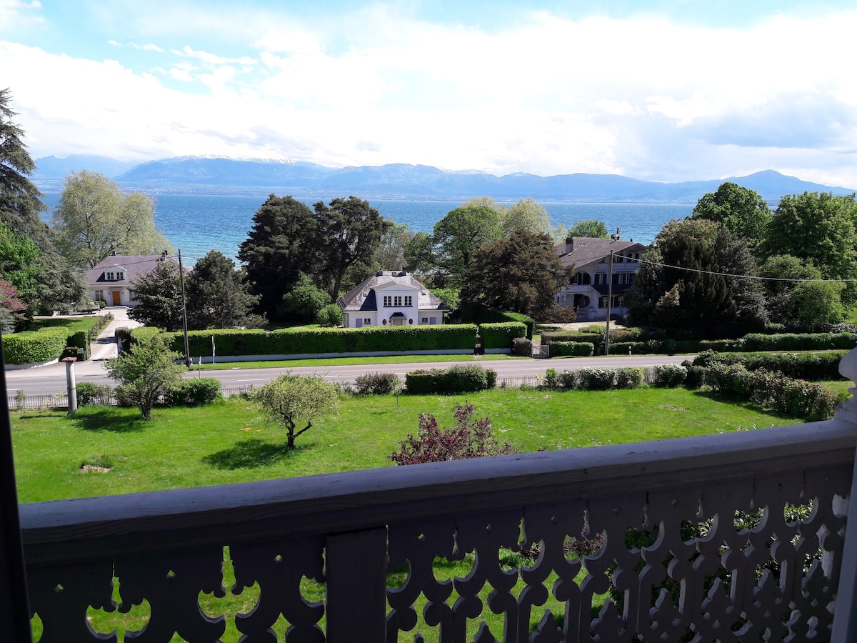 The image captures a scenic view from a balcony overlooking a lush green landscape. The foreground features well-manicured lawns and hedges, while the stunning Lake Geneva is visible in the background, framed by distant mountains under a bright sky.