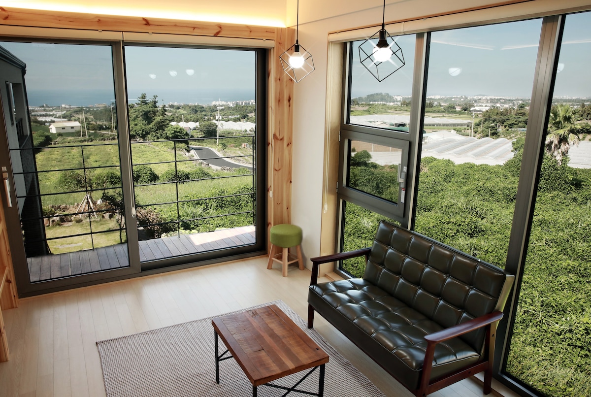 Bright, airy living space features large windows that frame a sweeping view of lush greenery and the distant coast. A compact wooden coffee table is centered in front of a dark, modern sofa. Natural light enhances the minimalist decor, creating a serene environment.