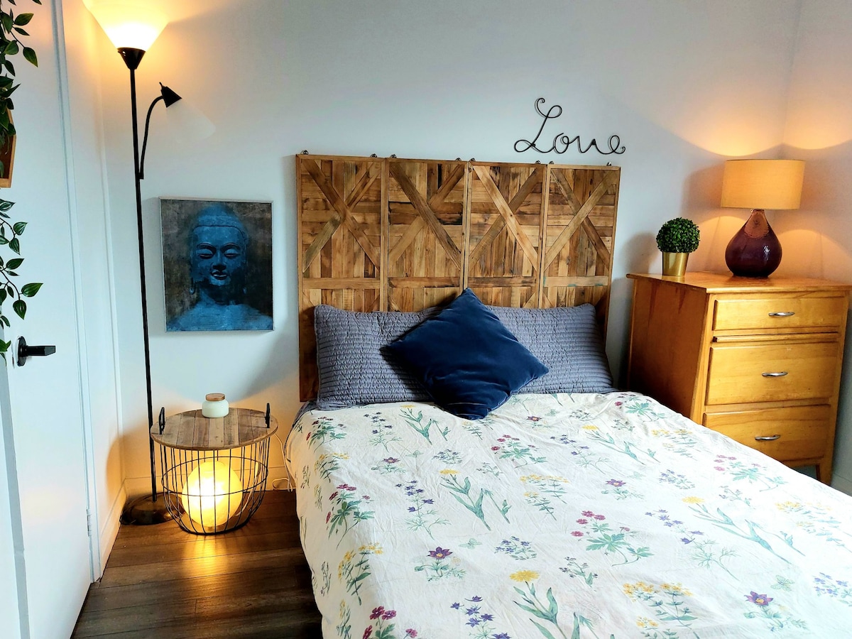 A serene bedroom space features a spacious bed with a floral-patterned coverlet and a large decorative cushion. A wooden headboard adds texture, while a warm lamp and a wooden nightstand provide soft lighting. A piece of art and a plant complete the inviting atmosphere.