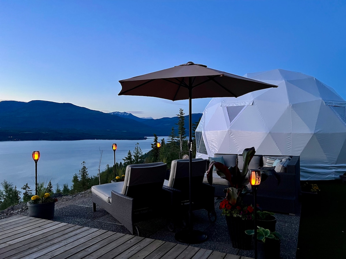 An outdoor seating area is situated in front of the geodesic dome, featuring two comfortable chairs and a table shaded by a large umbrella. Soft lighting from lanterns highlights the scenic backdrop of Shuswap Lake and the surrounding mountains as dusk approaches.