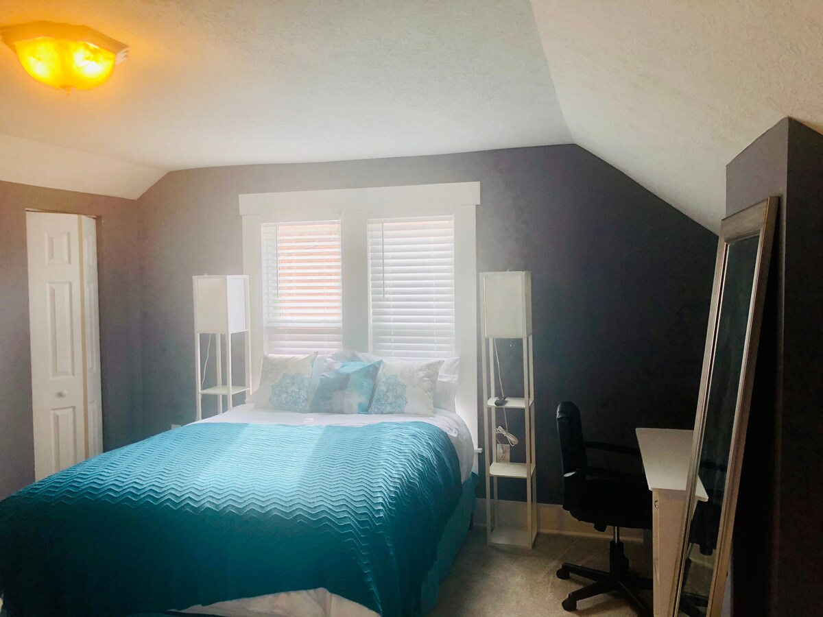 A cozy bedroom features a spacious queen bed with a textured blue blanket and decorative pillows. Natural light filters through two white-shuttered windows. A tall mirror and a desk with a chair are positioned near the bed, contributing to the room's functional layout.