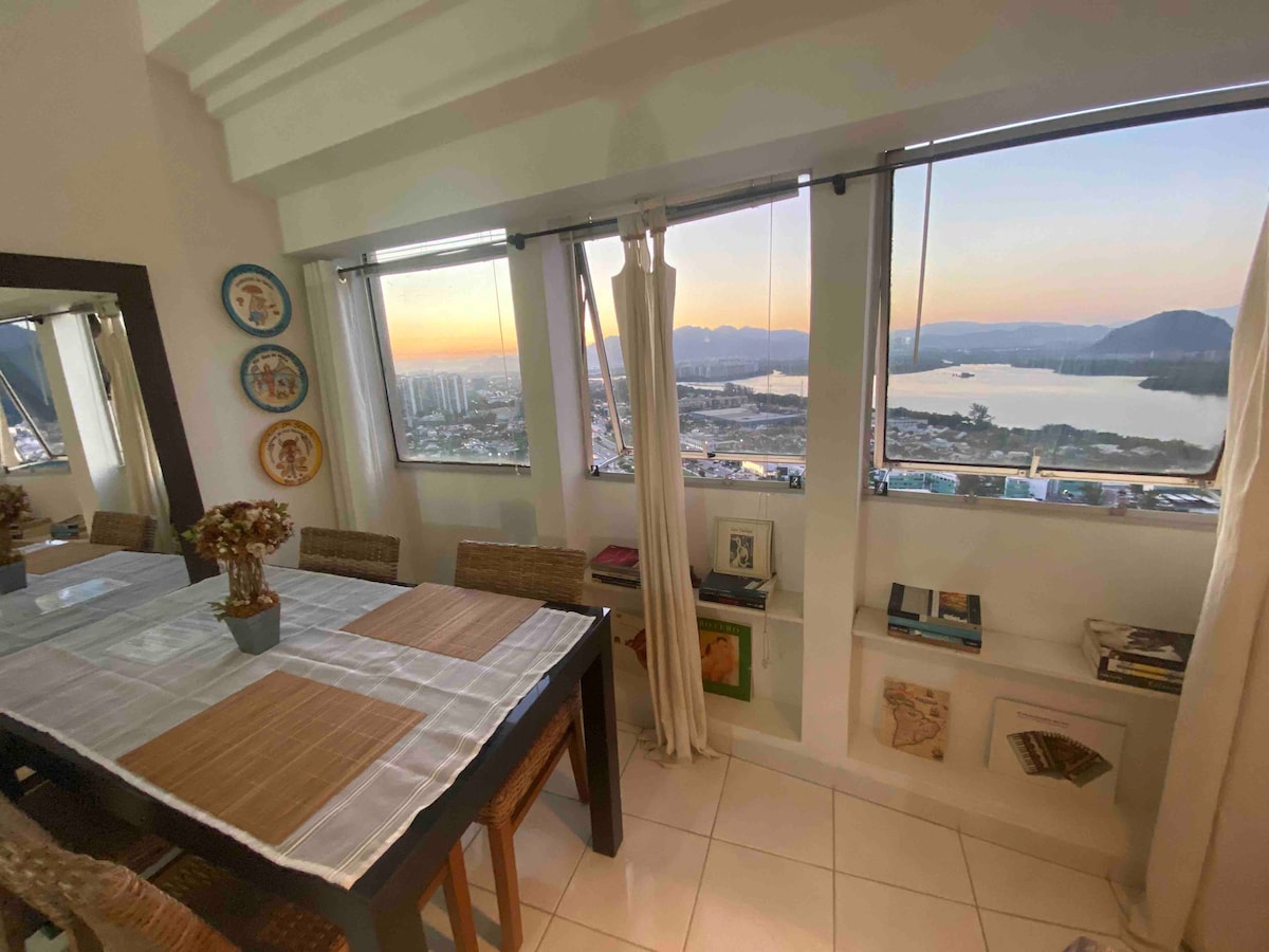 A dining area features a table that can accommodate four people. Natural light enters through large windows, offering a panoramic view of the surrounding landscape, including mountains and water. Decorative plates adorn the wall, enhancing the space's character.