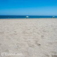 A sandy beach area is shown, leading towards the calm sea under a clear blue sky. Beach umbrellas are visible in the distance, adding a pop of color against the natural tones of the sand and water. The scene conveys a peaceful coastal atmosphere.
