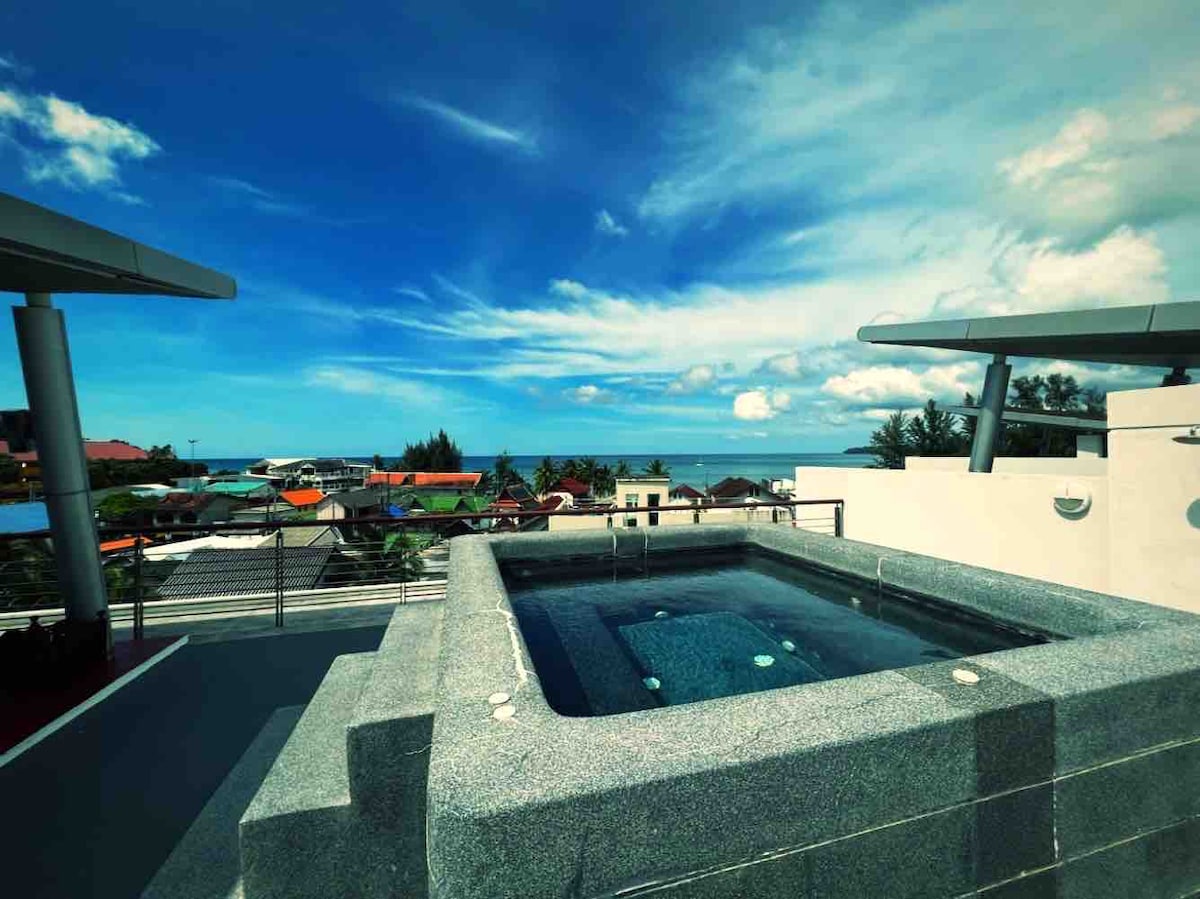 A private rooftop area features a spacious jacuzzi, surrounded by smooth stone walls. Panoramic views of the ocean and the vibrant landscape beneath are visible, with clear blue skies and scattered clouds overhead enhancing the inviting atmosphere.