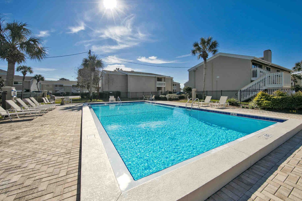 An inviting outdoor pool is framed by palm trees, with sun loungers arranged along the edge. The clear blue water reflects the bright sky and sunlight, creating a relaxing atmosphere for guests. Surrounding buildings are visible in the background.