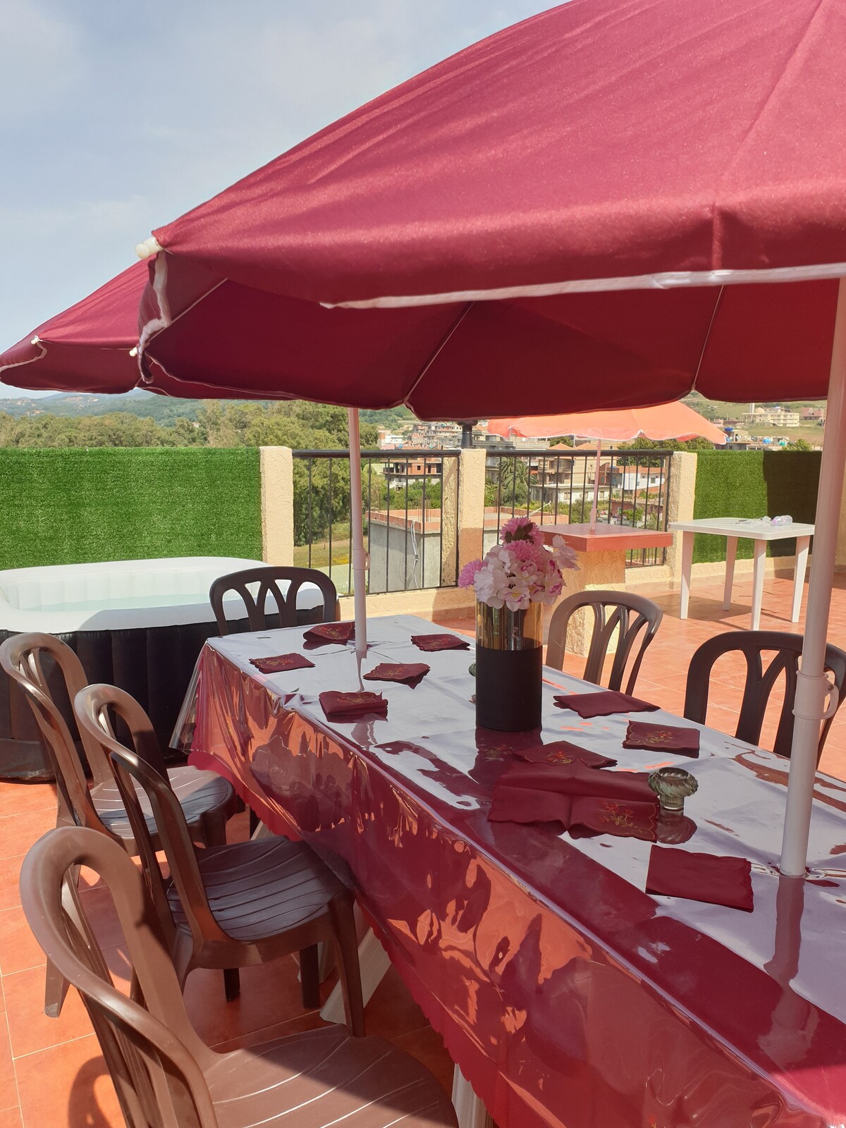 An outdoor dining area is presented under a large maroon umbrella. A long table, covered with a glossy maroon tablecloth, is set with neatly arranged napkins and a central vase holding flowers. Surrounding chairs are visible, with greenery and fencing enhancing the backdrop.
