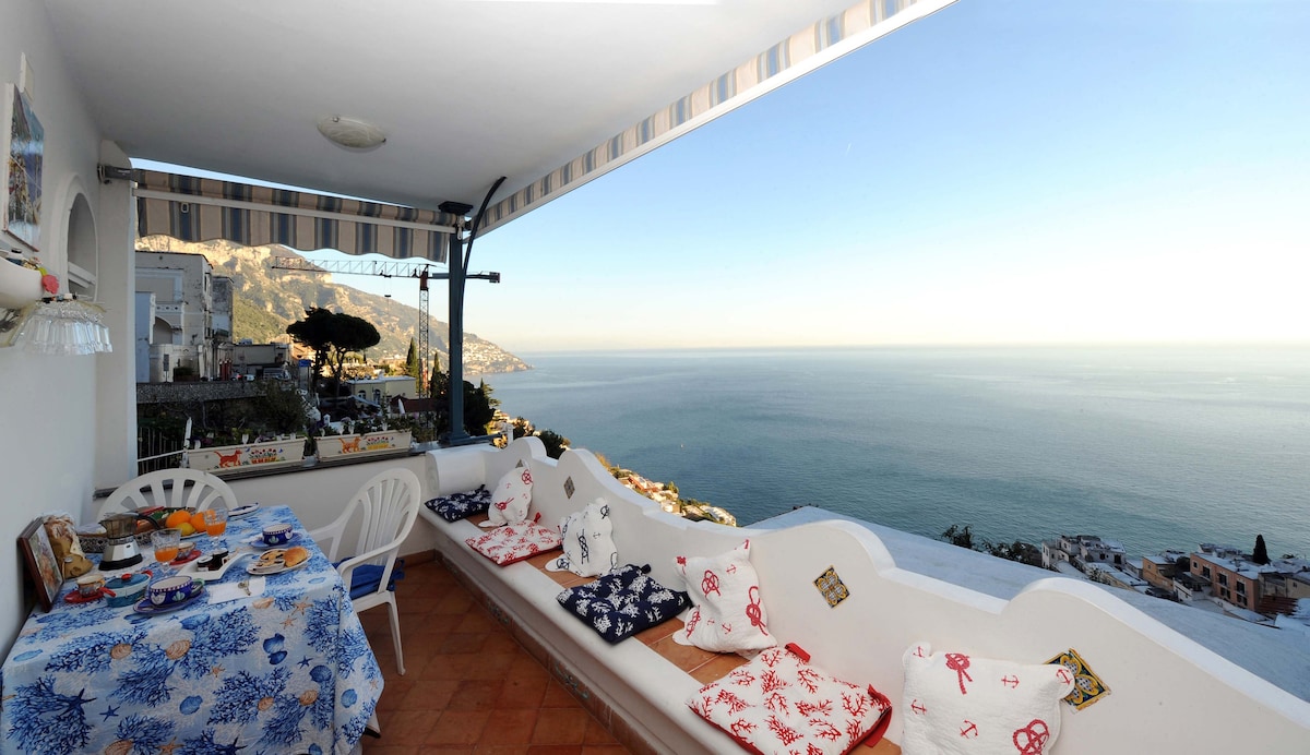 A private terrace is displayed, featuring a table set with breakfast items and a view of the sea. Comfortable seating with colorful cushions lines the edge, creating an inviting space to enjoy the coastal scenery.