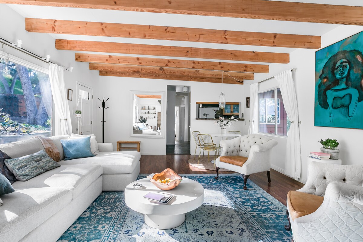 A bright living area features exposed wood beams and large windows allowing natural light to fill the space. A white sectional sofa and an elegant tufted chair are arranged around a circular coffee table atop an ornate area rug, complemented by artwork on the wall.