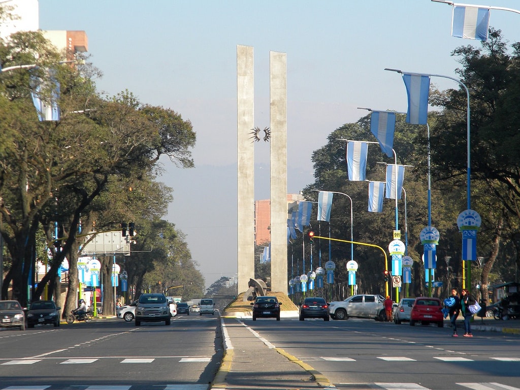 The image displays a wide avenue lined with large trees and decorative blue and white banners. At the center stands a tall monument with two vertical columns, flanked by vehicles on the road and pedestrians on the sidewalk.