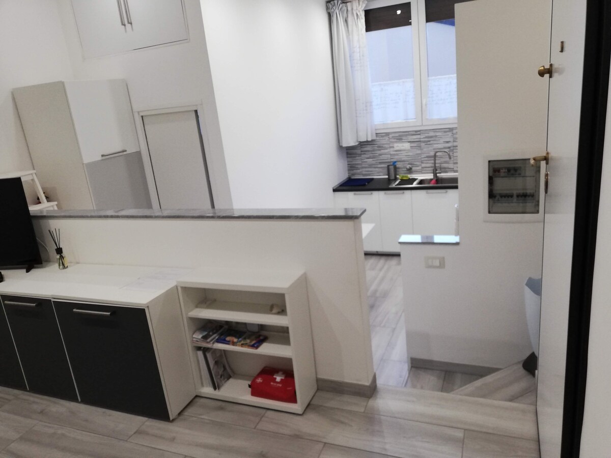 The living area features a modern open-plan layout with light-colored cabinetry and a spacious countertop. A small dining table is visible along with kitchen areas, while natural light filters through sheer curtains at the window. A staircase leads to the lower level.