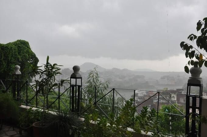 A scenic view of the landscape is captured from a balcony, with lush greenery framing the foreground. The clouds are overcast, hinting at rain, while distant hills and buildings are visible through the mist. The railing is adorned with ornamental posts, enhancing the scene.