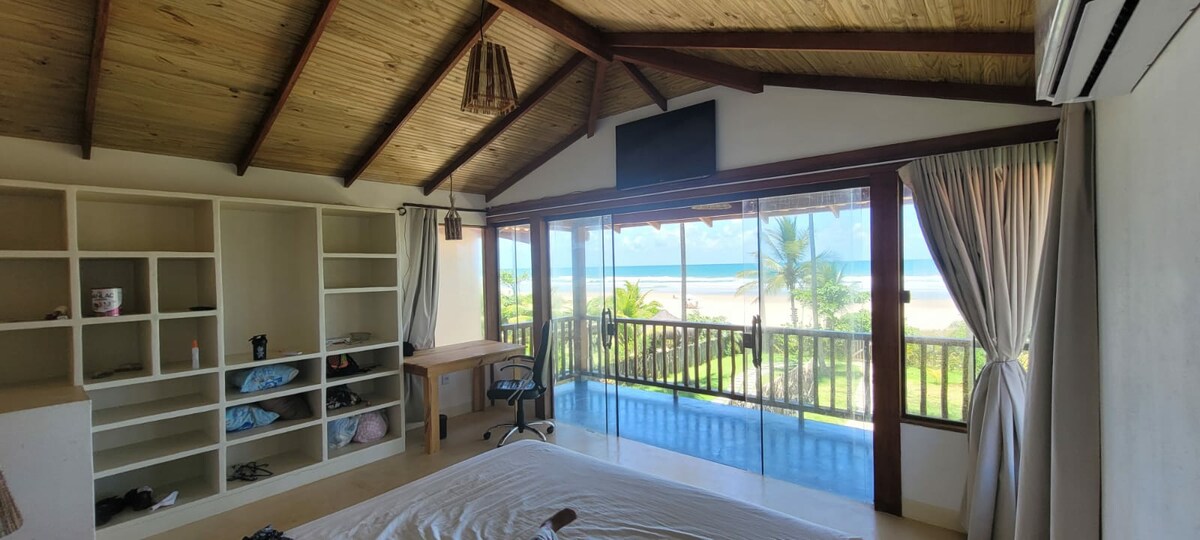 A spacious bedroom features high wooden ceilings and large windows offering ocean views. A comfortable bed is positioned centrally, with a work desk and chair nearby. Light curtains frame the windows, allowing natural light to fill the room. Shelving is visible for storage.