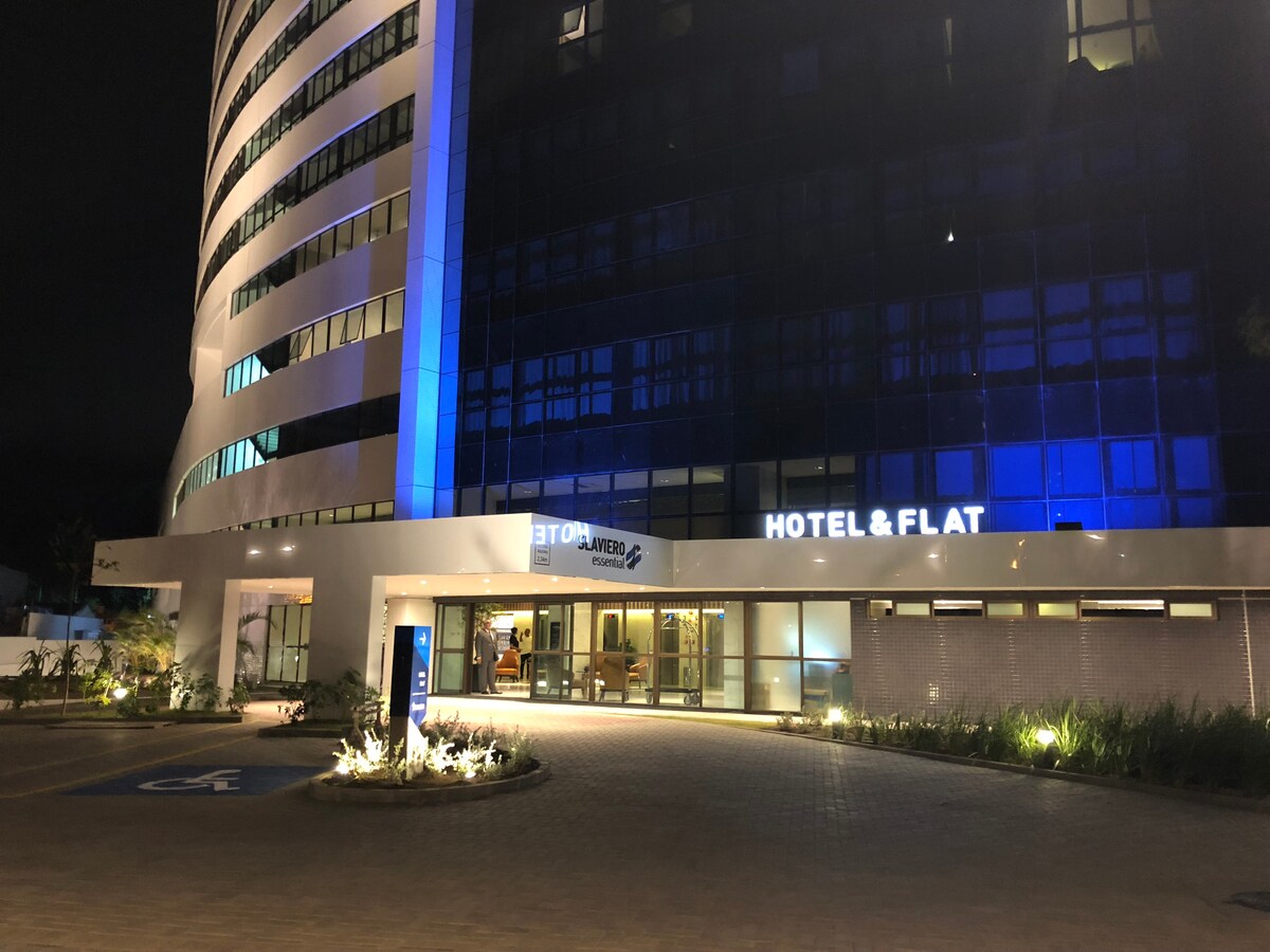 The hotel facade is illuminated in blue, featuring modern architecture with large glass windows. The entryway is highlighted by a welcoming atmosphere, displaying the hotel name prominently. Surrounding landscaping includes subtle lighting, adding to the inviting appearance of the complex at night.