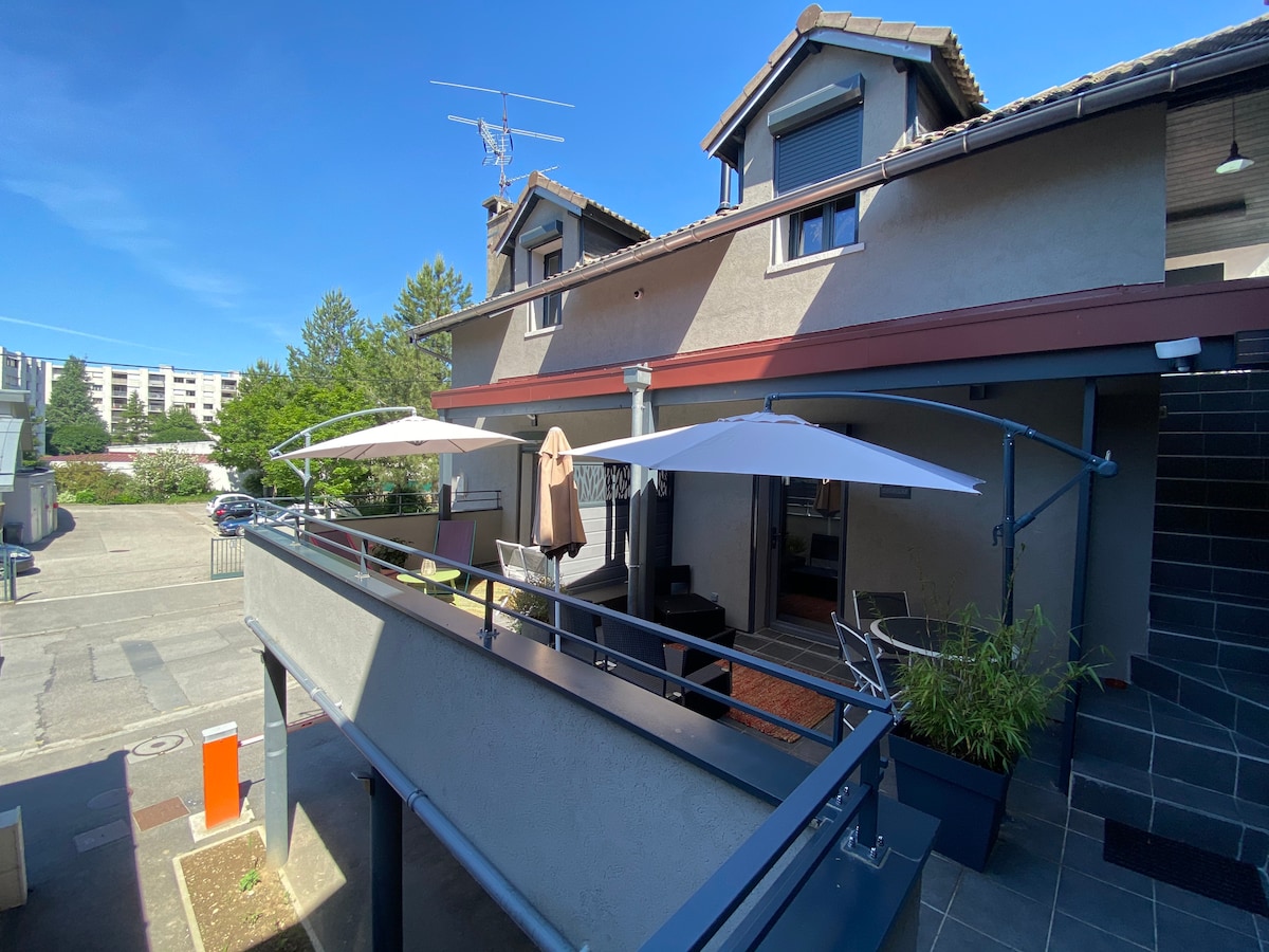 An outdoor terrace is showcased with two large umbrellas providing shade over seating arrangements. The building’s upper level is visible, featuring a modern structure with large windows. A sense of calm is conveyed through the surrounding greenery and the quiet street below.
