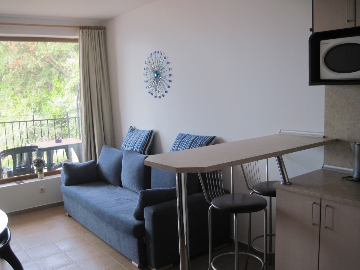 A comfortable living area features a dark blue sofa with coordinating cushions, positioned near a large window. A compact dining counter with two bar stools complements the space, while light cream walls enhance the room's brightness. Outdoor views of greenery are visible through the window.