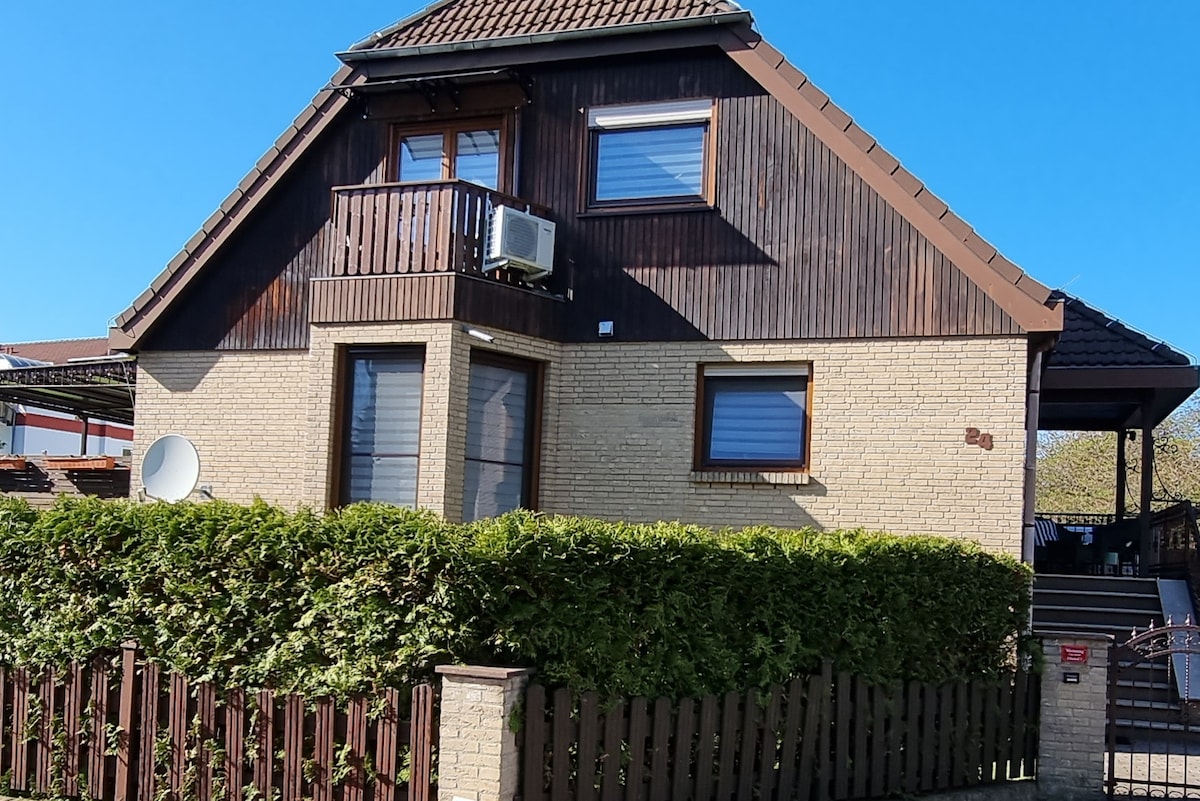 A charming two-story house features a mix of brick and wooden accents. Large windows provide natural light, while a small balcony offers outdoor space. The property is framed by a low wooden fence and greenery, creating a welcoming appearance. An air conditioning unit is visible on the exterior.