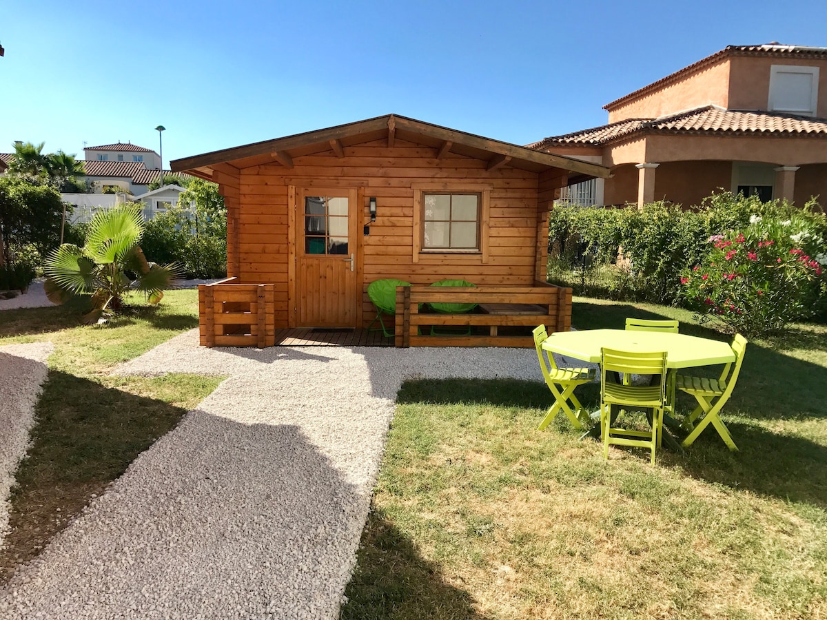 A charming wooden chalet is set against a clear blue sky, surrounded by lush greenery. A gravel path leads to the chalet, which features a front porch and a seating area. Bright green outdoor furniture complements the inviting landscape.