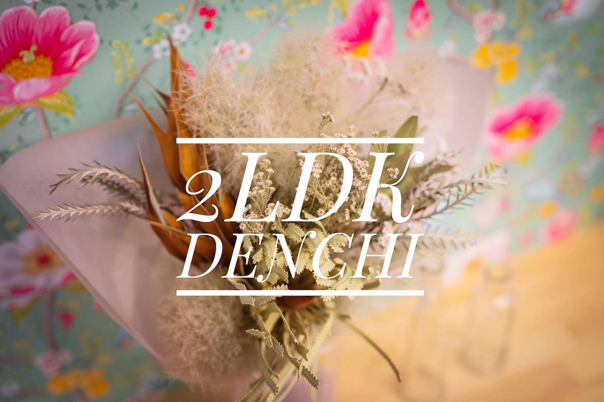 A floral arrangement featuring dried flowers and greenery is presented against a colorful floral-patterned wallpaper. The arrangement combines various textures, adding a natural touch to the decor. Soft lighting enhances the subtle colors of the flowers in this inviting setting.